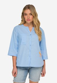 Jasnoniebieska koszula z guzikiem, z kieszonką, 3/4 rękawami i pomarańczowym nadrukiem po bokach. Wykonana z gładkiego, teksturowanego materiału.