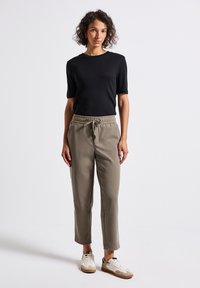 Vrouw met krullend haar staat gekleed in een zwart shirt met korte mouwen, grijze broek met koord en witte sneakers tegen een effen witte achtergrond.