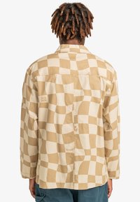 Kevyt, pitkähihaisella puserolla beigen sävyssä, jossa on aaltoileva ruutukuosi. Varustettu klassisella kauluksella ja näkyvillä ompelutiedoilla selässä.