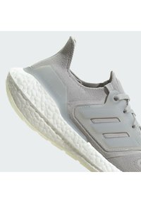 Scarpa da corsa grigio chiaro con una parte superiore testurizzata, dettagli in rete e una suola bianca imbottita. Presenta tre strisce parallele sul lato.