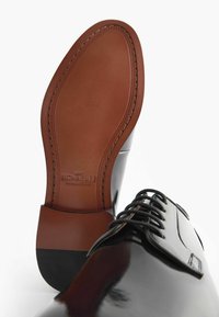 Chaussure habillée en cuir noir avec une finition lisse, bout rond et lacets noirs. La semelle en cuir marron présente des coutures visibles et un logo.
