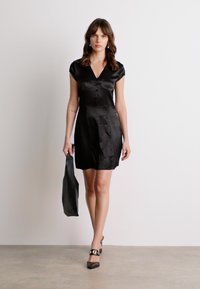 Robe en satin noir avec des manches courtes, un décolleté en V et des détails de boutons. Le mannequin porte un sac noir assorti et des chaussures à talons noires avec des accents ronds.