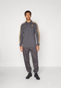 Grå pikétröja med lång ärm och två knappar i sprundet, guldfärgade ränder på ärmarna, broderad logga, matchad med gråa joggerbyxor och svarta skor.