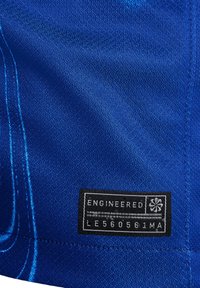 Sininen teksturoitu kangas, jossa on etiketti, jossa lukee "ENGINEERED", sekä suunnitteluelementti. Näkyvää ompelua ja materiaalin kuvioiden vaihtelua.