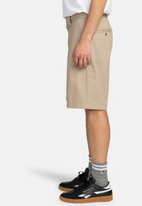 Pantaloni corti beige alla lunghezza del ginocchio realizzati in cotone, con design frontale piatto, tasche laterali e passanti per cintura, abbinati a sneakers nere e calzini grigi a righe.