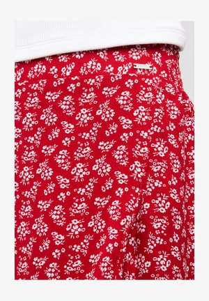 Czerwony materiał z małym białym wzorem kwiatowym, z dekoracyjną metką logo. Materiał wydaje się lekki i ma gładką fakturę.