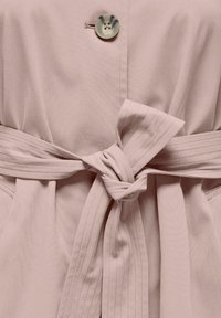 Robe portefeuille rose clair avec une ceinture nouée, dotée d'un unique bouton rond et d'une texture de tissu lisse. Plis discrets à la taille.