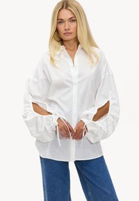 Chemise blanche à manches ballons, avec des découpes au niveau des coudes et des fermetures à cordon. Tissu lisse et coupe décontractée.