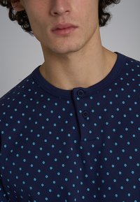 Camicia button-up blu navy con piccoli motivi a cerchi verdi, scollatura arrotondata e placket con due bottoni. Tessuto morbido con una texture liscia.