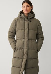 Frau in einem olivgrünen, langen gepolsterten Wintermantel mit Kapuze, mit Händen in den Taschen, steht vor einem schlichten hellen Hintergrund.
