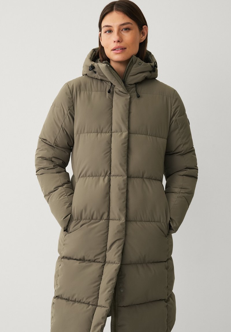 Frau in einem olivgrünen, langen gepolsterten Wintermantel mit Kapuze, mit Händen in den Taschen, steht vor einem schlichten hellen Hintergrund.