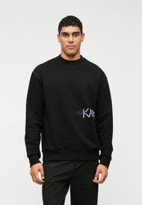 Ung man i svart sweatshirt med blå och vit text på framsidan, stående mot en enkel ljus bakgrund.