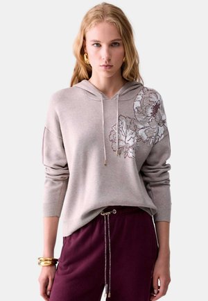 Donna bionda che indossa una felpa beige con cappuccio e ricamo floreale sulla spalla, abbinata a pantaloni viola scuro con coulisse e gioielli in oro.