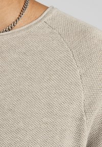 Zbliżenie na beżowy sweter dzianinowy w okolicach ramienia i dekoltu z srebrnym naszyjnikiem na skórze.