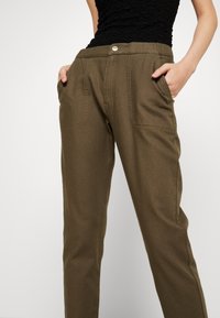 Olijfgroene broek met twee voorzakken, een normale tailleband en een knoopsluiting. Gemaakt van een gestructureerde stof met zichtbare stiksels.