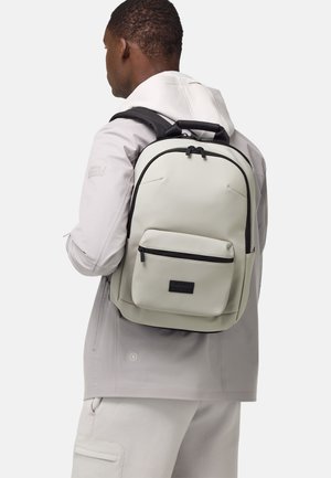 Zaino grigio chiaro con dettagli neri, dotato di una tasca anteriore con zip, spallacci regolabili e un design elegante e arrotondato.