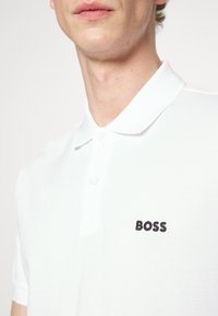 Vit kortärmad polo med en strukturerad design, som har två knappar och en svart broderad "BOSS"-logotyp på bröstet.