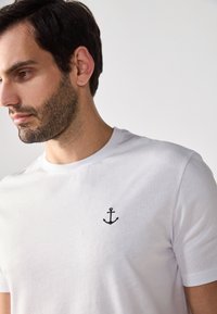 Hvid bomulds t-shirt med en sort broderet anker på venstre bryst. Rund hals og korte ærmer. Glat tekstur.
