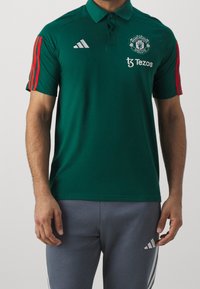 Zelené polo tričko vyrobené z hladké látky, s límečkem, tříknoflíkovou légou, červenými pruhovanými akcenty a logem Manchester United.