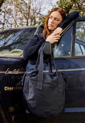Denim-Schultertasche mit gerafftem oberen Teil, dunkelblauer Farbe, flachem Boden und stabilen Tragen. Enthält ein dezent gehaltenes Markenlogo in Kupfer.