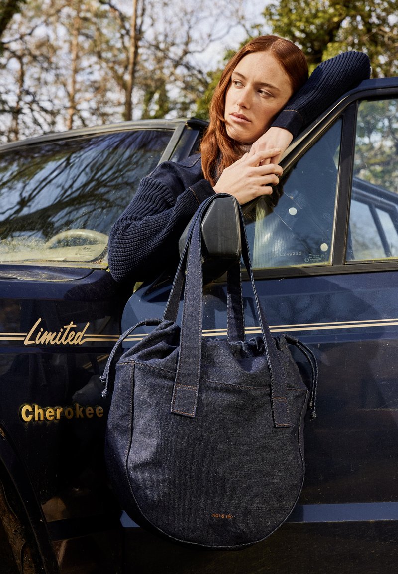 Sac en denim avec une fermeture coulissante sur le dessus, de couleur bleu foncé, fond plat et anses robustes. Présente un logo de marque subtil en cuivre.