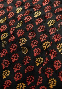 Tissu noir présentant un motif floral et de feuilles répétitif en rouge, jaune et or. Le design a une apparence texturée avec des formes distinctes.