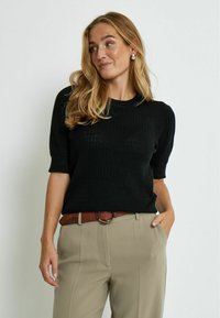 Pull noir en tricot avec des manches courtes bouffantes et un col rond, associé à un pantalon beige taille haute et une ceinture marron avec une boucle en métal.