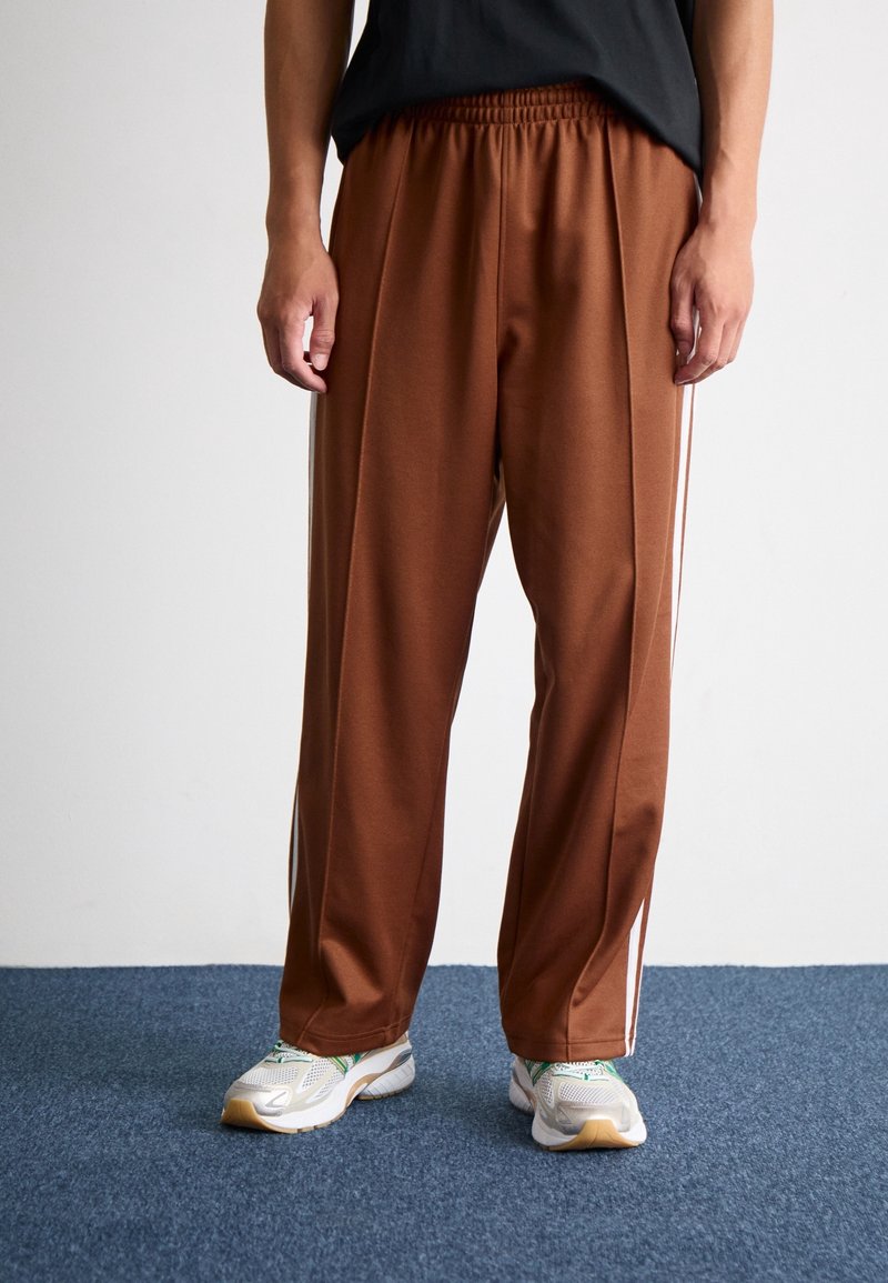 Braune Sporthose mit elastischem Bund, weißen Seitenstreifen und lockerer Passform, kombiniert mit grau-grünen Turnschuhen auf einem blauen Teppich.