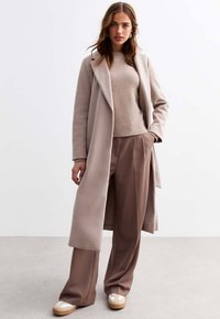 Manteau long beige avec un col à revers, associé à un pull en tricot et un pantalon large marron ; baskets claires avec une semelle plateforme.
