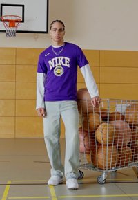 T-shirt violet Nike avec des manches blanches, jean bleu clair, baskets blanches, se tenant à côté d'un chariot à fils rempli de ballons de basket dans un gymnase.