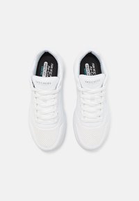 Des chaussures de sport blanches présentent un matériau synthétique, des détails perforés, une forme arrondie, des lacets plats et une semelle intérieure rembourrée avec un logo sur la languette.