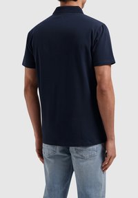 Marineblauwe poloshirt met een gestructureerde kraag, korte mouwen en een subtiele logodetail op de rug. Gemonteerd met lichtblauwe jeans.