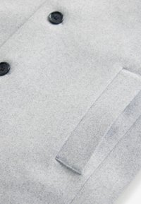 Abrigo de lana gris claro con una superficie suave y texturada, que presenta dos botones negros y un bolsillo único. Diseño minimalista con líneas limpias.