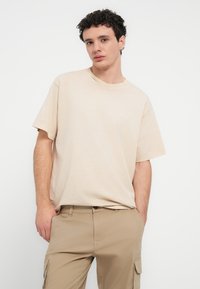 Μπεζ βαμβακερό T-shirt με κοντά μανίκια και στρογγυλή λαιμόκοψη. Υφασμα με υφή και λεπτομέρεια με διακριτικό λογότυπο στο στήθος. Συνδυασμένο με χακί παντελόνια.