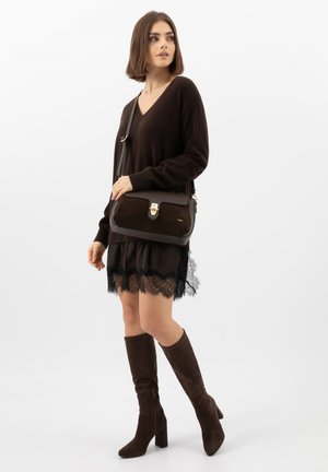 Žena v tmavě hnědém svetru s výstřihem do V, černé sukni s krajkovým lemem, hnědých kozačkách po kolena a s hnědou koženou kabelkou přes rameno.