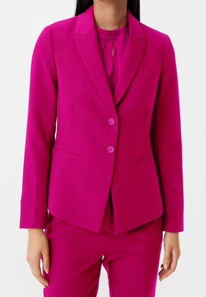 Kvinde iført en skarpt magenta skræddersyet blazer med to knapper og matchende bukser mod en ensfarvet lys baggrund.