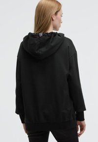 Black hoodie made of soft fabric, featuring a drawstring hood, dropped shoulders, and ribbed cuffs and hem. Back includes a small label.