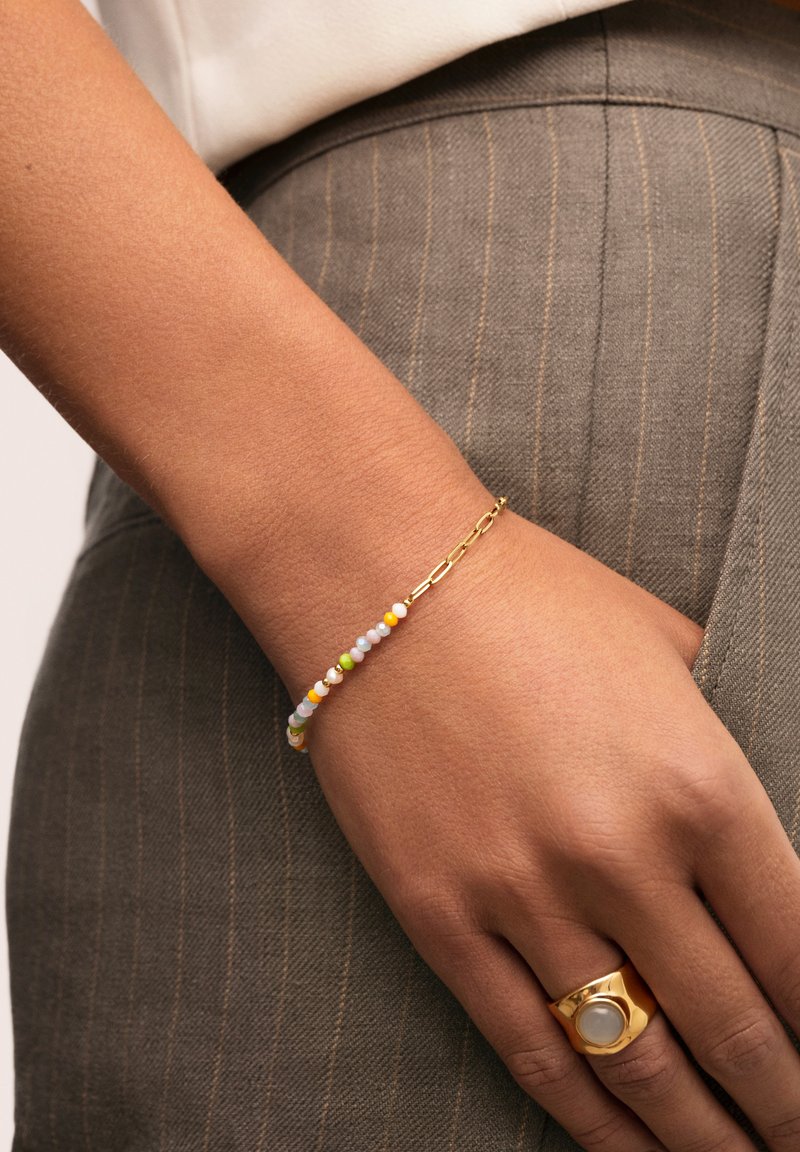 Goldene Kettenarmband mit mehrfarbigen Perlen in Pastellfarben, getragen an einem Handgelenk, kombiniert mit einem goldenen Ring und grauem Nadelstreifenstoff.