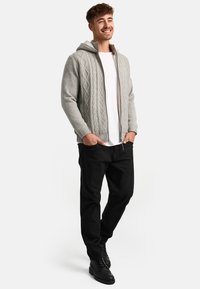 Sudadera con cremallera de punto en gris claro con un patrón de trenzas, combinada con una camiseta blanca y pantalones negros. La sudadera cuenta con capucha y puños acanalados.