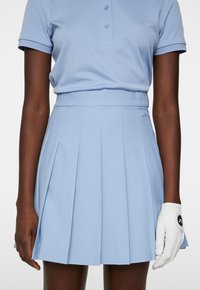 Polo golf azzurro chiaro con patta a tre bottoni, abbinato a una gonna plissettata coordinata. Una guanto da golf bianco è indossato sulla mano sinistra.