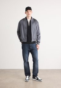 Chaqueta bomber gris sobre una sudadera negra con cremallera, combinada con jeans azul oscuro y zapatillas negras y blancas. El modelo lleva una gorra negra.