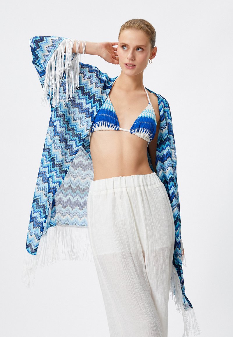 Blauwe en witte zigzag patroon kimono met franjes, gedragen over een blauw en witte bikini en witte luchtige broeken.