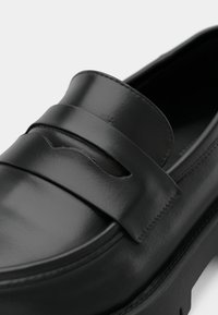 Musta nahkainen loafer-kenkä kiiltävällä viimeistelyllä, jossa on paksu remmi ja katkaistuja yksityiskohtia. Tasainen pohja takaa helpon käytön.