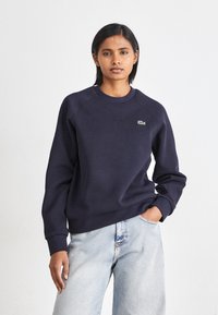 Marinegenser med rund hals, ribbestrikkede mansjetter og en liten grønn logo på brystet. Stoffet ser mykt og strukturert ut. Brukt med lyseblå jeans.