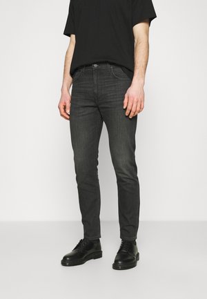 Man wearing black slim-fit jeans, black t-shirt, and black lace-up shoes, standing against plain light background.