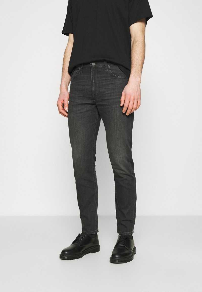 Homme portant un jean noir slim, un t-shirt noir et des chaussures à lacets noires, debout devant un fond clair uni.