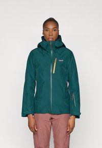 Teal waterdichte jack met capuchon, volledige rits aan de voorkant, gele ritszak en verstelbare manchetten. Draagt over lichtroze broek.