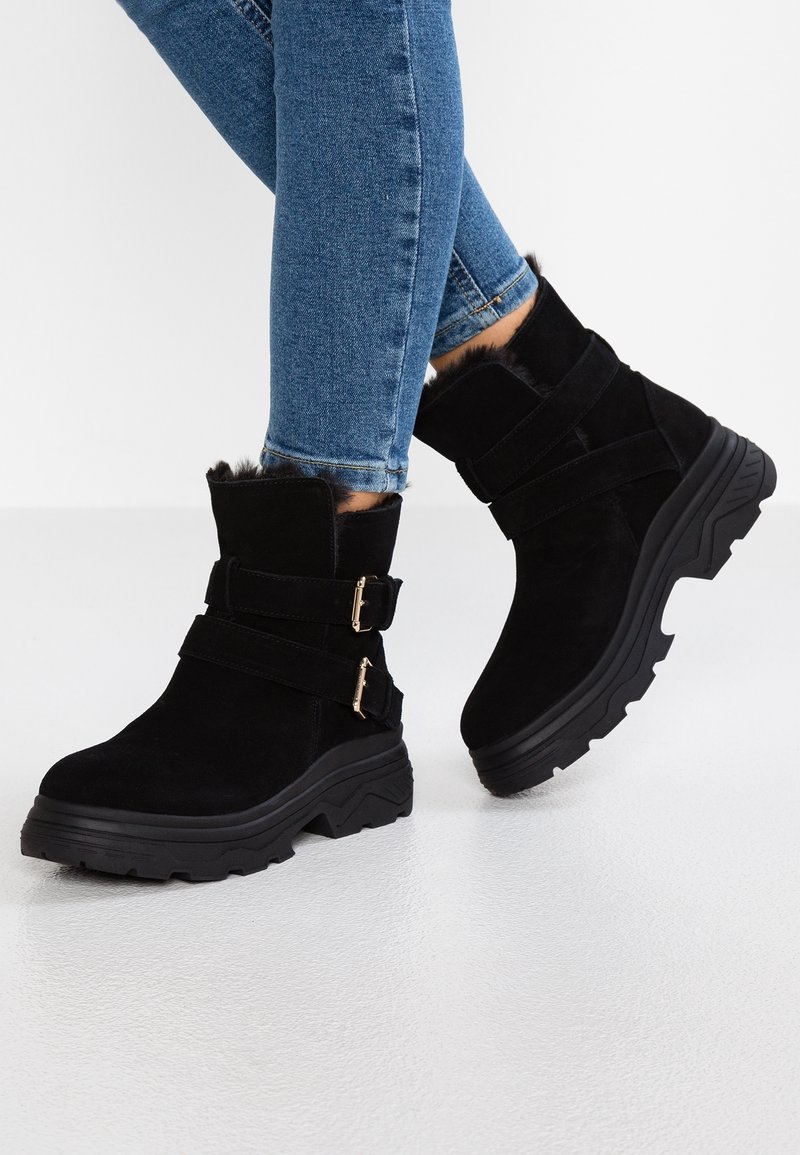 Botas de tobillo de ante negro con ribete de pelo sintético, diseño de doble correa con hebillas metálicas y suela gruesa negra, combinadas con vaqueros azules.