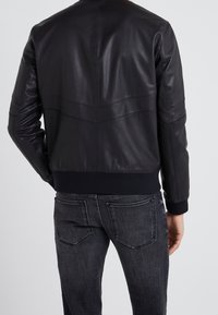 Czarna skórzana kurtka bomberka z prążkowanym kołnierzem i mankietami, ozdobiona pionowymi przeszyciami na panelach oraz dopasowanym paskiem w talii, zestawiona z ciemnoszarymi dżinsami.