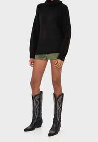 Schwarzer Strickpullover mit hohem Kragen, kombiniert mit grünen Shorts. Tragend mit schwarzen Leder-Cowboystiefeln mit silberner Stickerei.